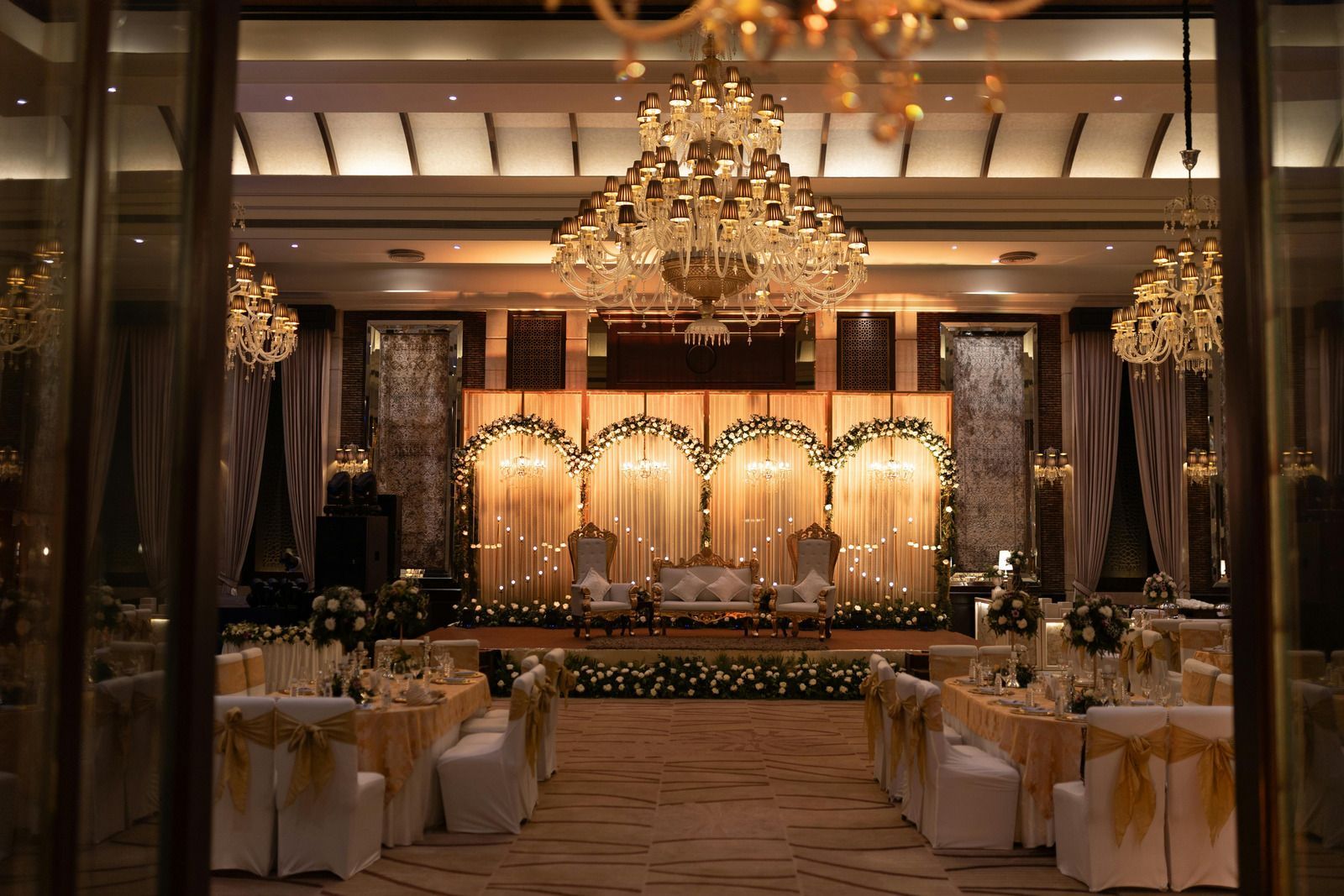 Elegant banquet hall with chandeliers, set tables, and a lit stage at the far end