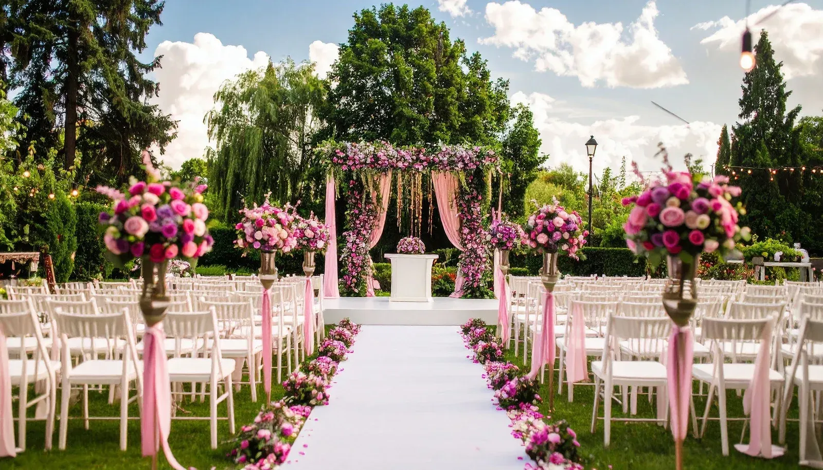 White wedding aisle lined with pink flowers and chairs under a floral arch outdoors