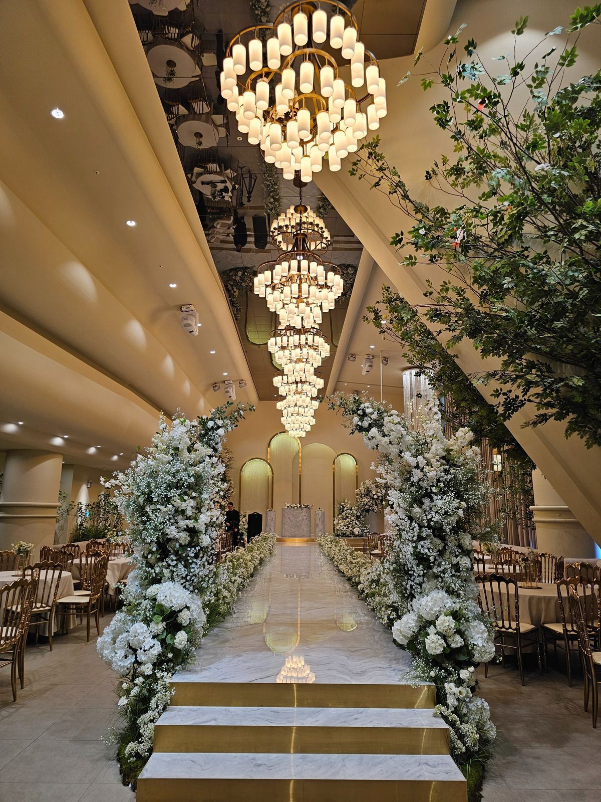 Elegant wedding aisle with white floral arrangements, chandelier lights, and gold chairs in a grand hall