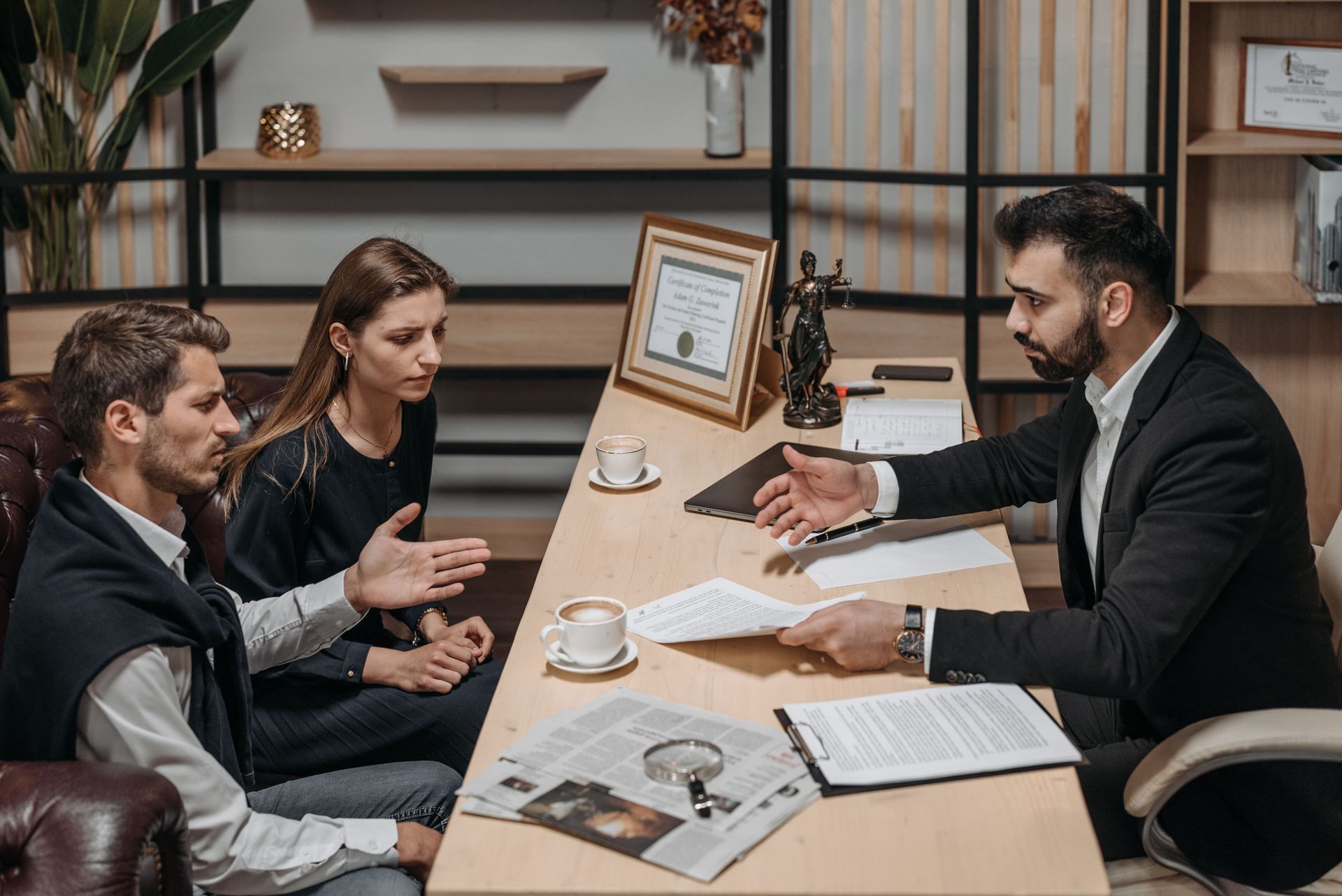 Two people meet with a suited advisor at a conference table, reviewing documents in a modern office.