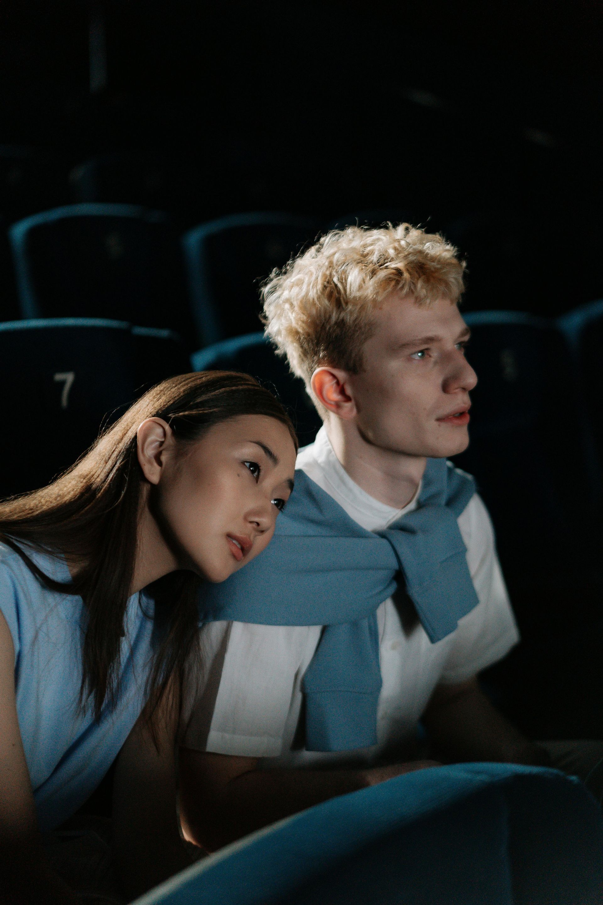 Two people sit in a dark theater, watching a film, with blue sweater draped over their shoulders.
