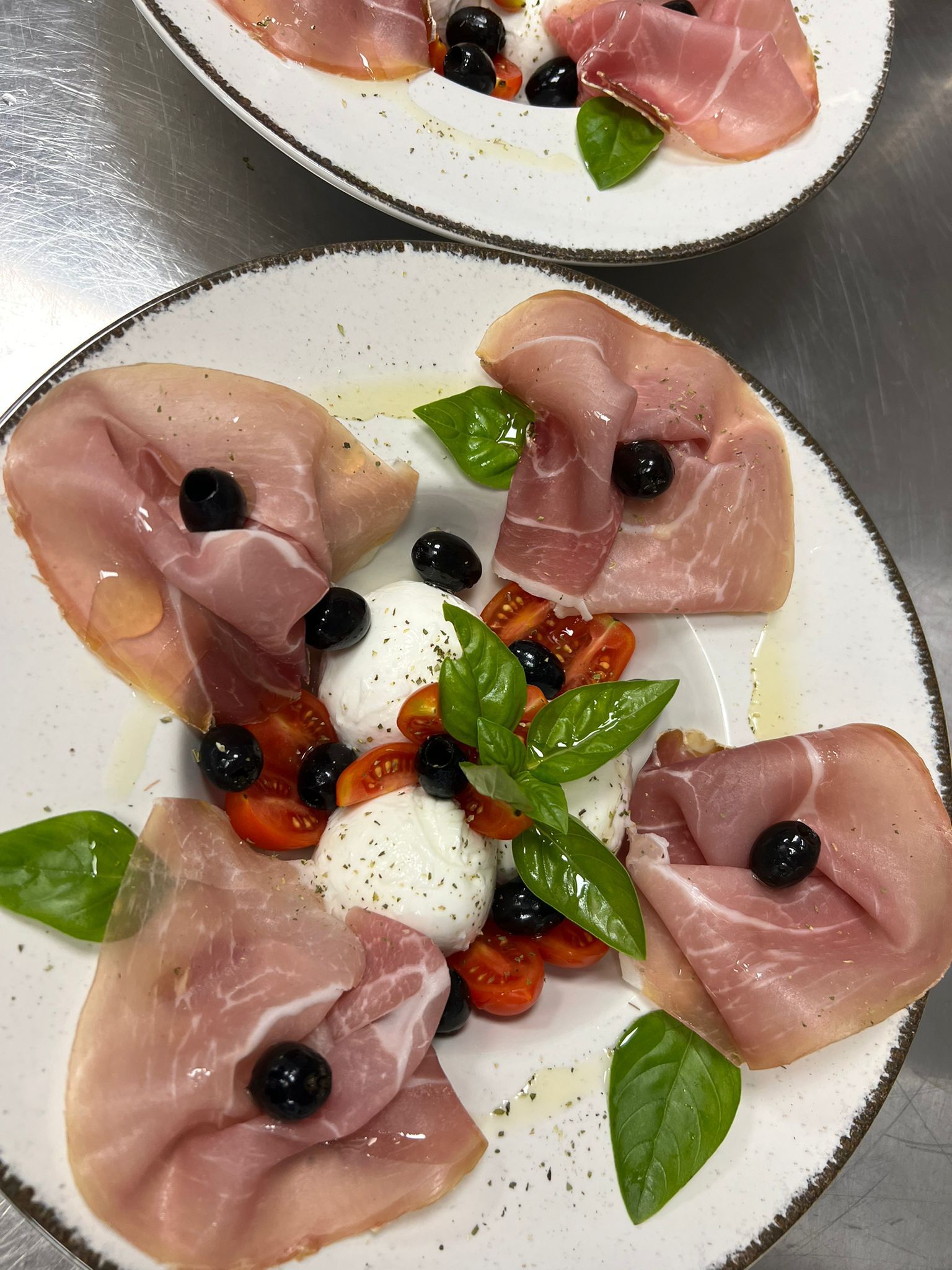 Melone avvolto nel prosciutto crudo, con palline di mozzarella, foglie di basilico, olive e pomodorini, servito su un piatto bianco.