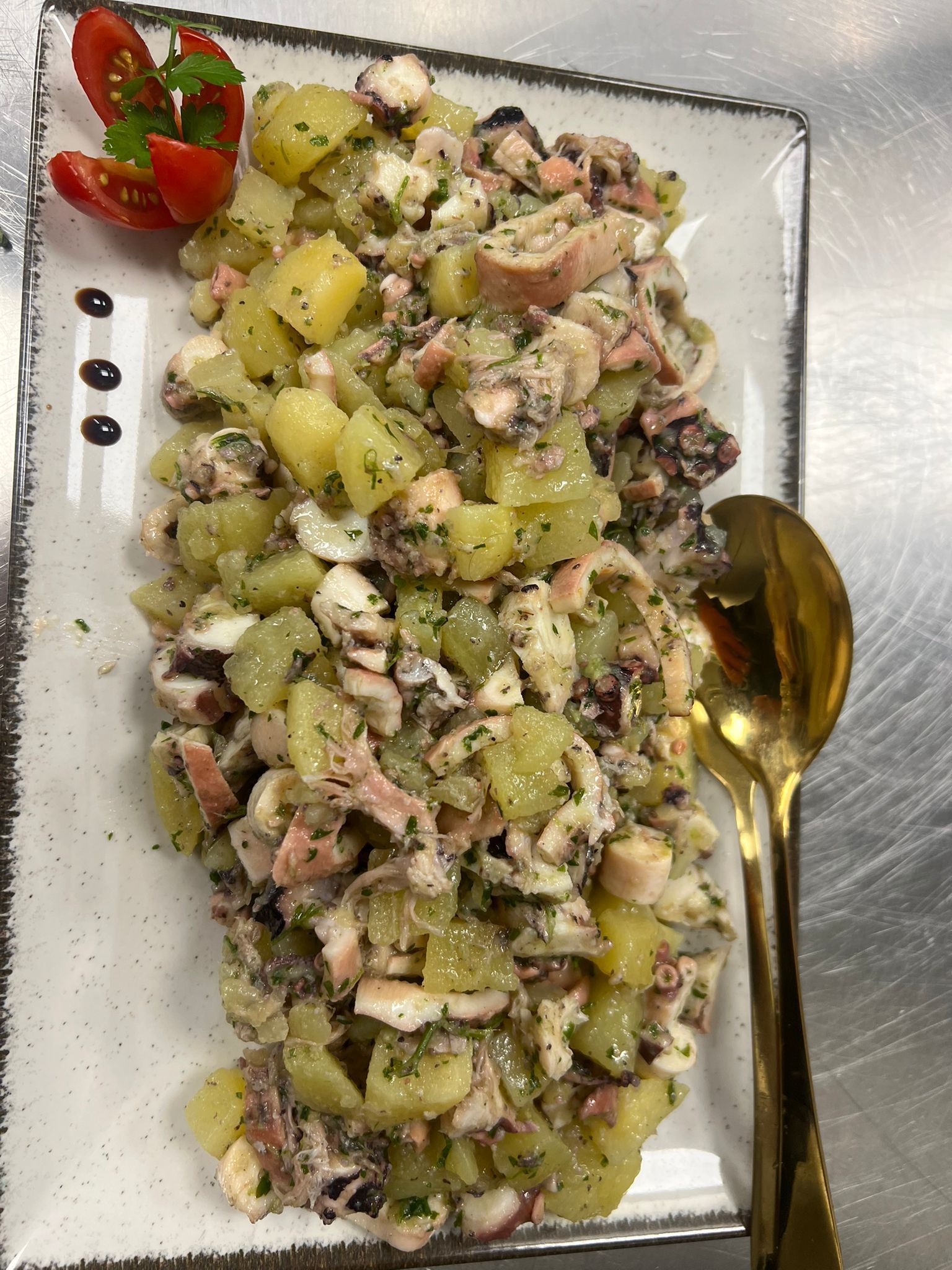 Insalata di patate con erbe aromatiche e tonno su un piatto bianco, con pomodorini e un cucchiaio dorato.