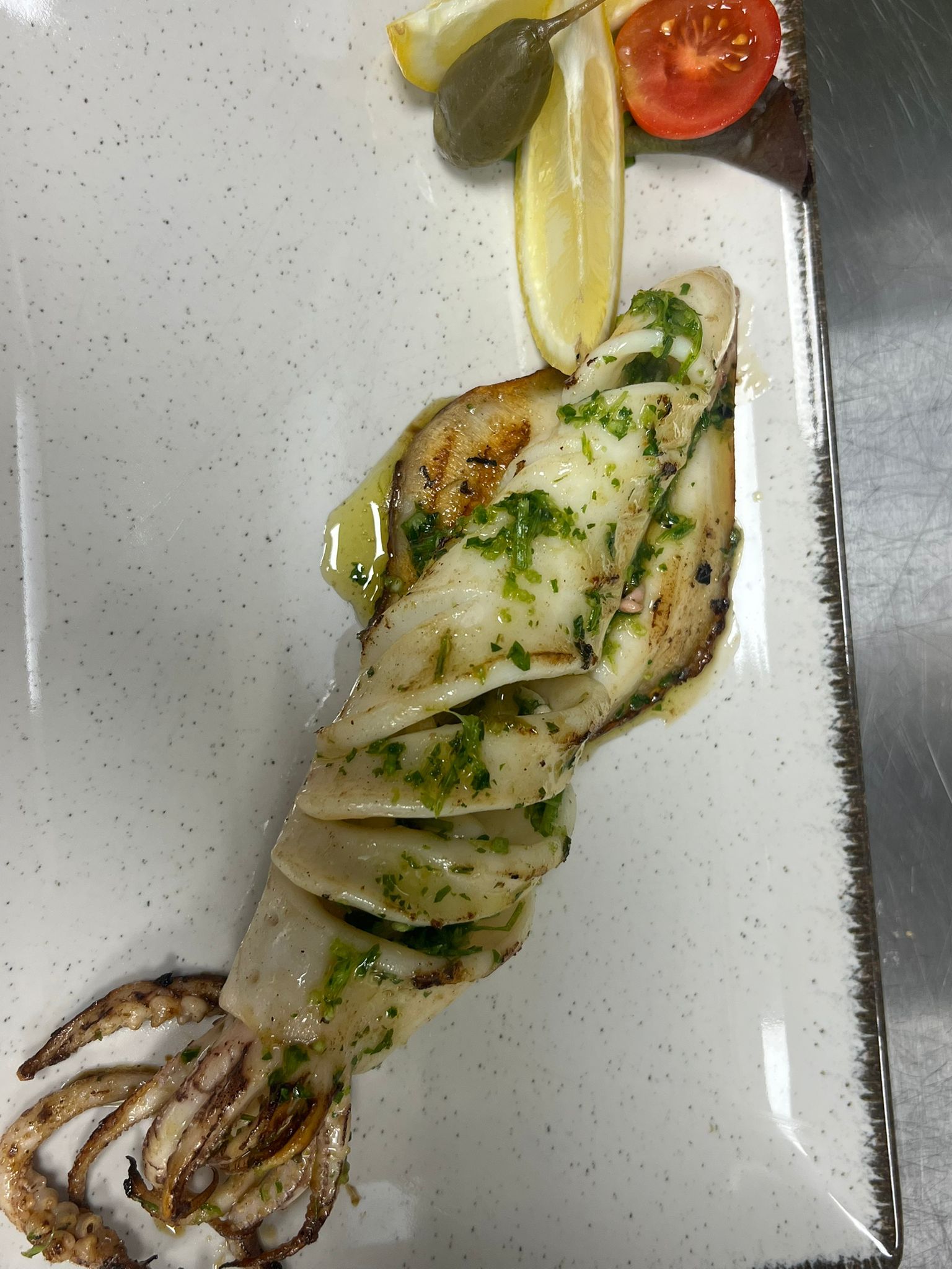 Calamari grigliati su un piatto bianco con un filo di erbe aromatiche e una guarnizione di pomodorini ciliegino.