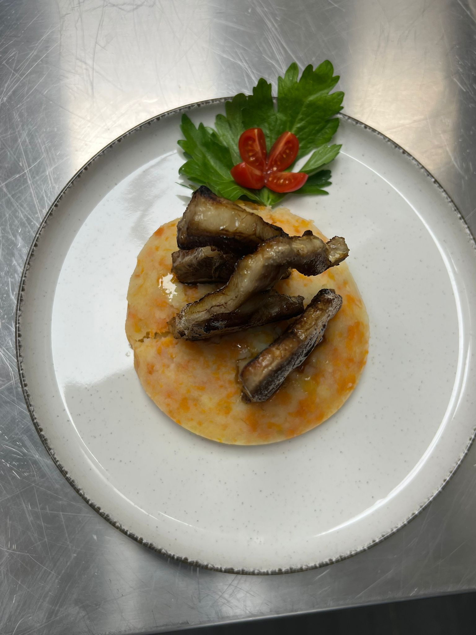 Piatto di pesce impiattato su un piatto bianco con prezzemolo, pomodorini e salsa all'arancia su una superficie in acciaio inox.