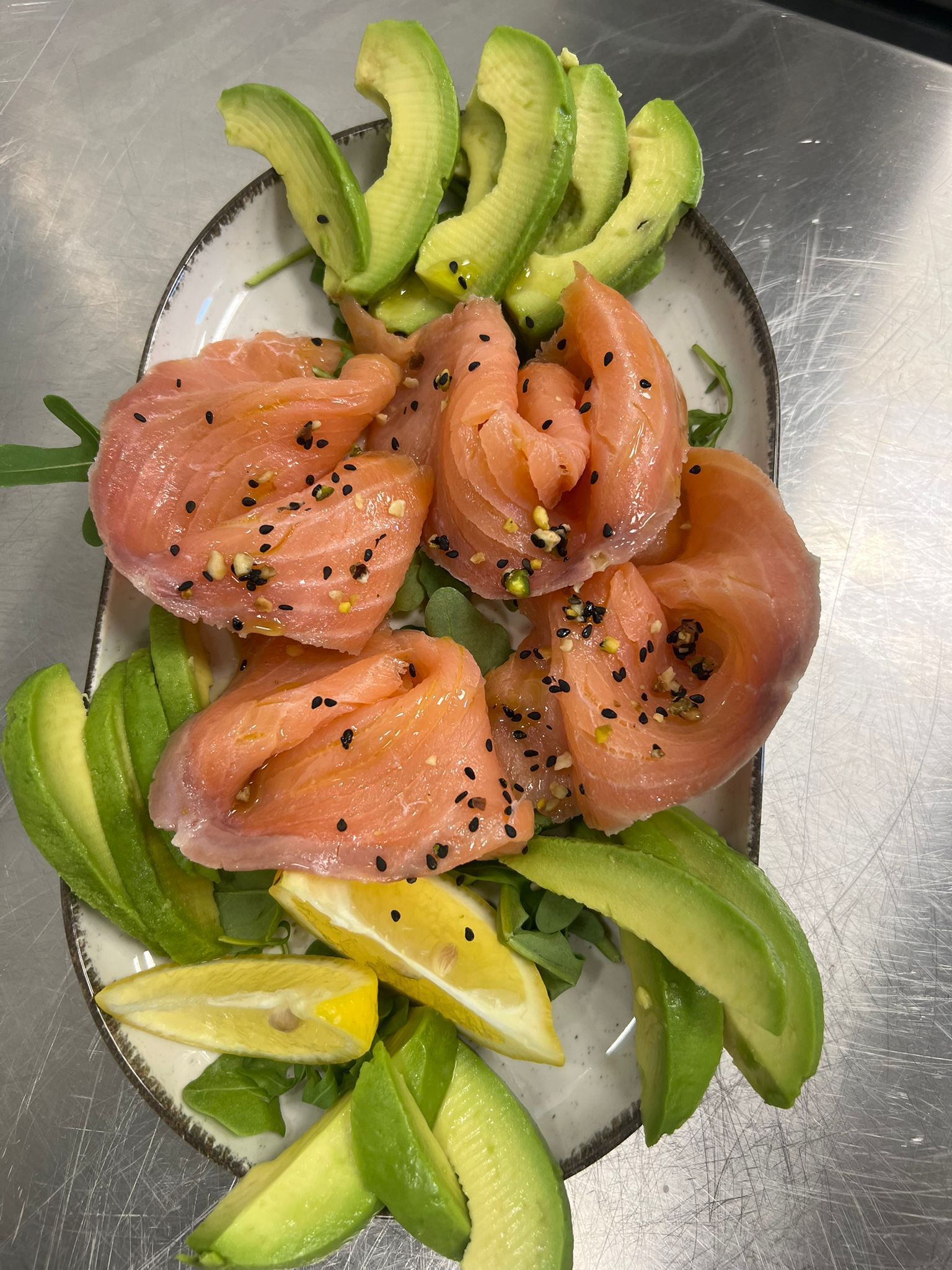 Sashimi di salmone disposto su un piatto di metallo con fette di avocado e spicchi di limone, guarnito con semi di sesamo.