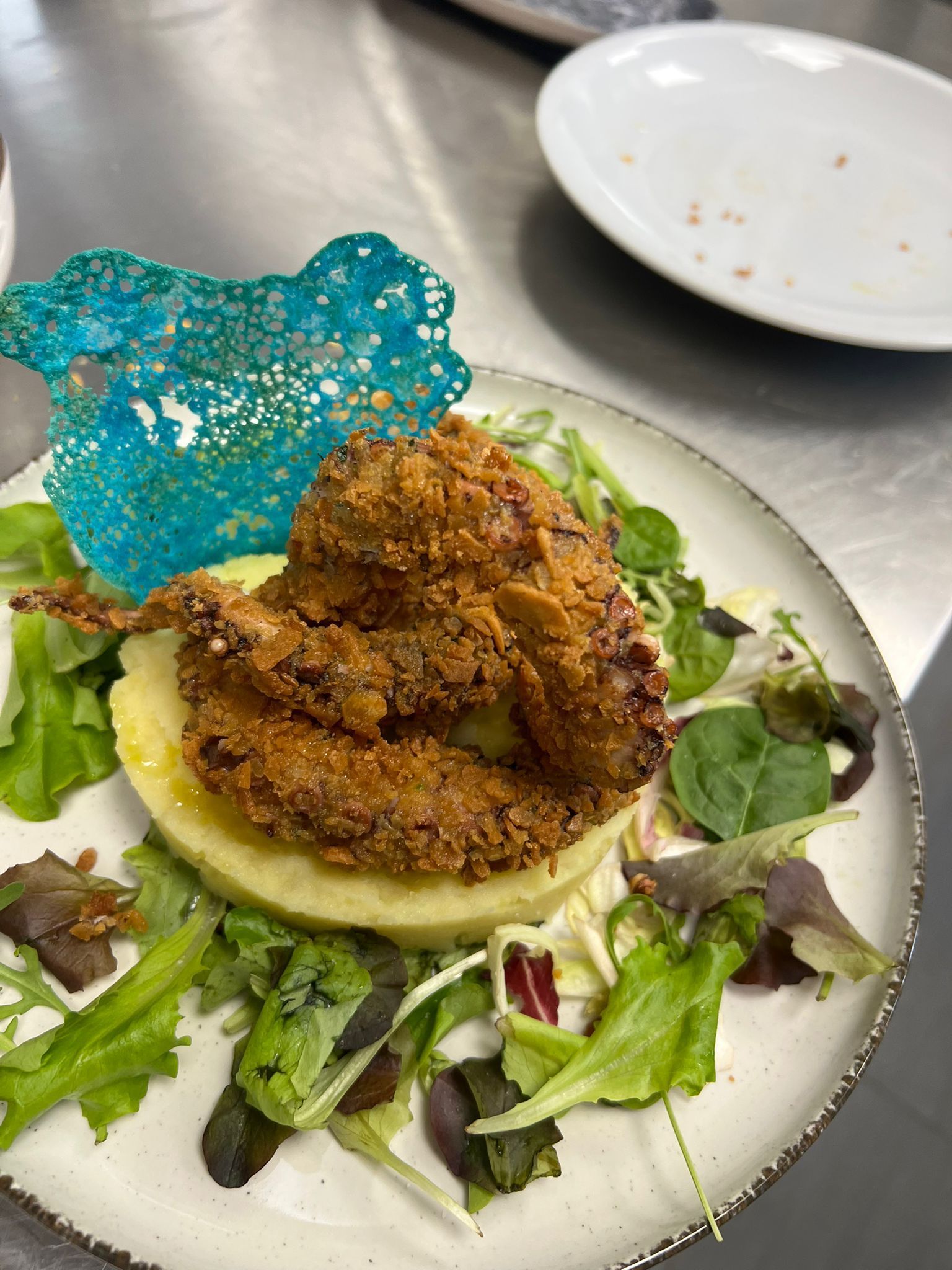 Insalata con guarnizione fritta su una fetta di limone, guarnita con una cialda di pizzo blu, su un piatto bianco.