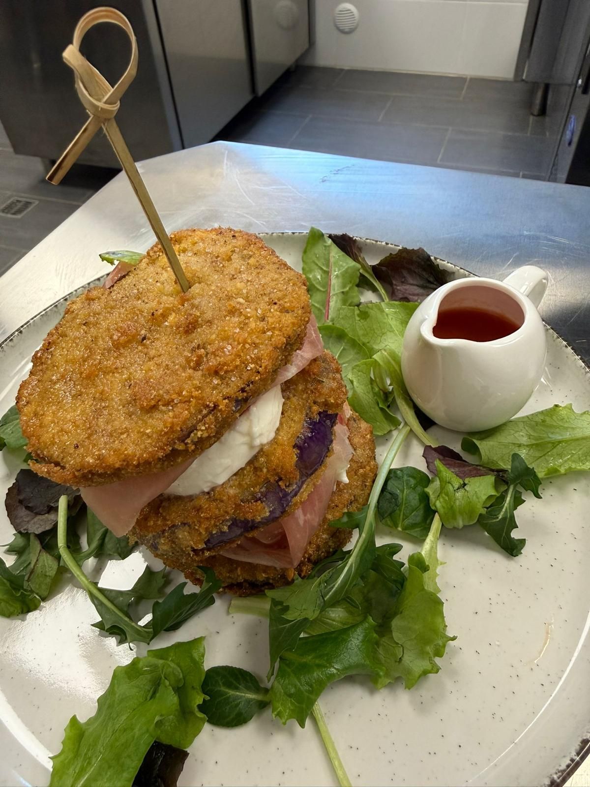 Panino gourmet con una crosticina croccante su un piatto bianco, guarnito con verdure e una ciotolina di salsa rossa.