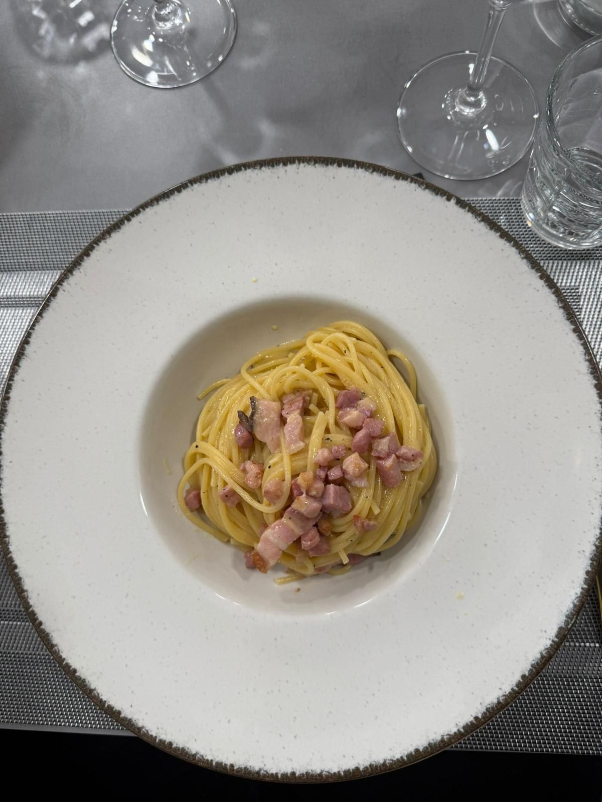 Piatto di spaghetti alla carbonara con pancetta in una ciotola bianca su un tavolo apparecchiato con bicchieri da vino.