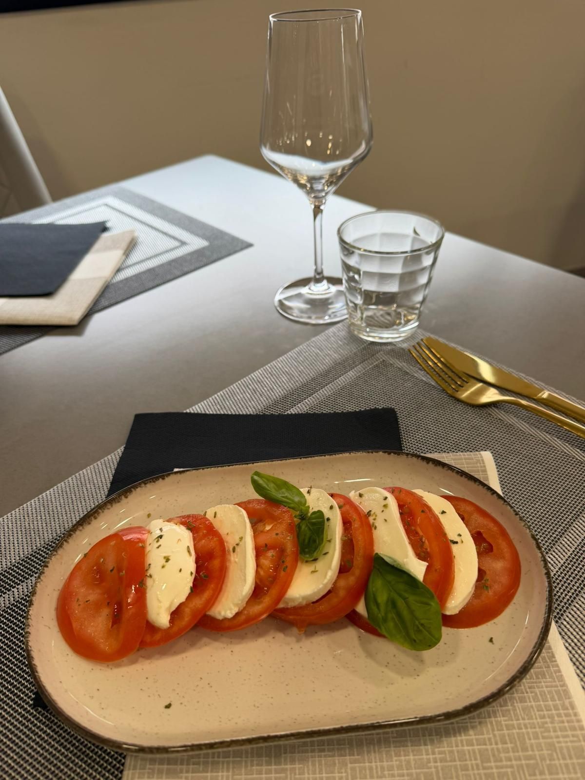 Insalata Caprese su un piatto con pomodoro, mozzarella, basilico, e un bicchiere da vino e un bicchiere d'acqua sul tavolo.