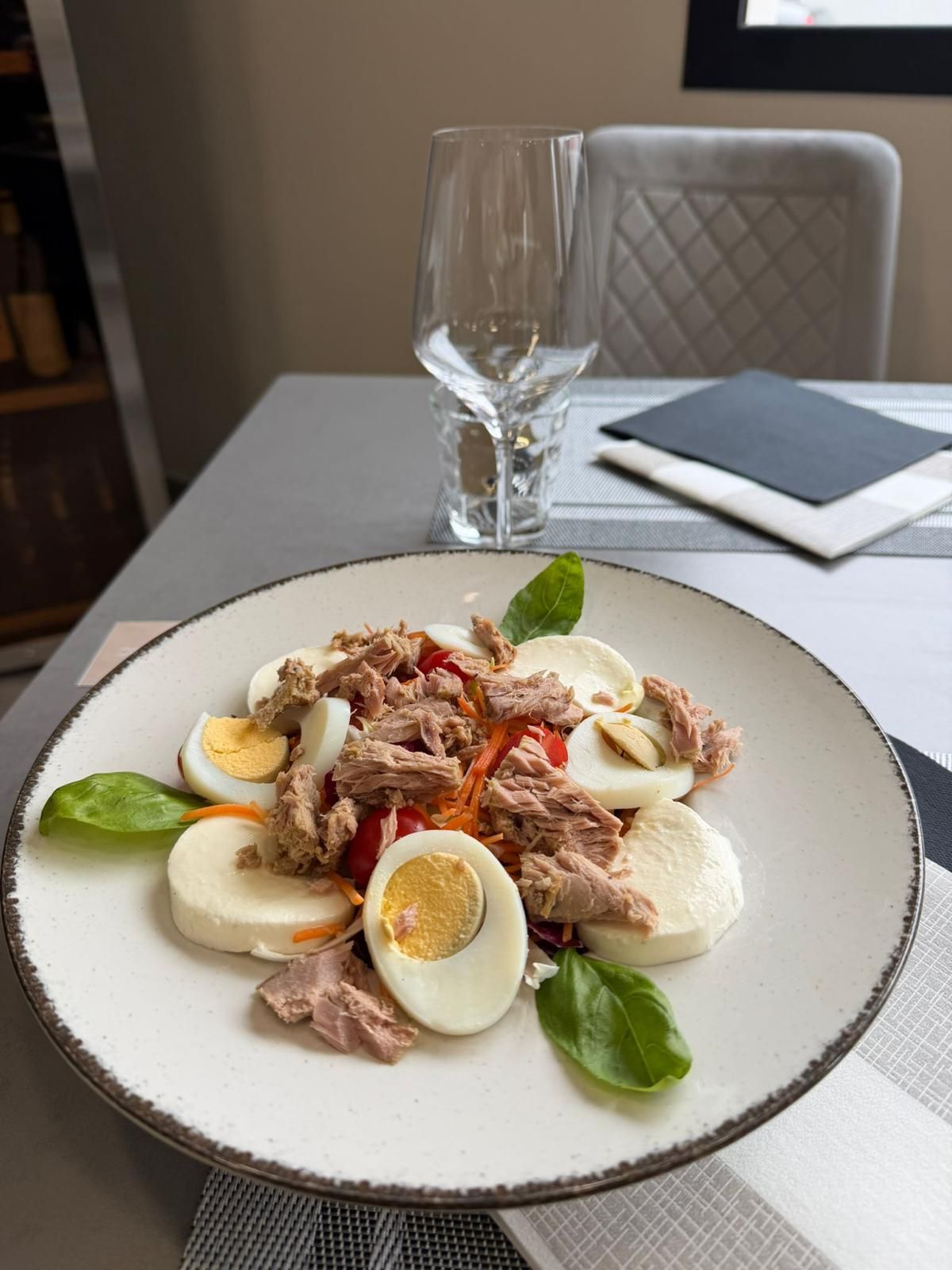 Un piatto di insalata di tonno con uova sode e basilico su un tavolo da pranzo, con un bicchiere di vino vuoto e un tovagliolo nelle vicinanze.