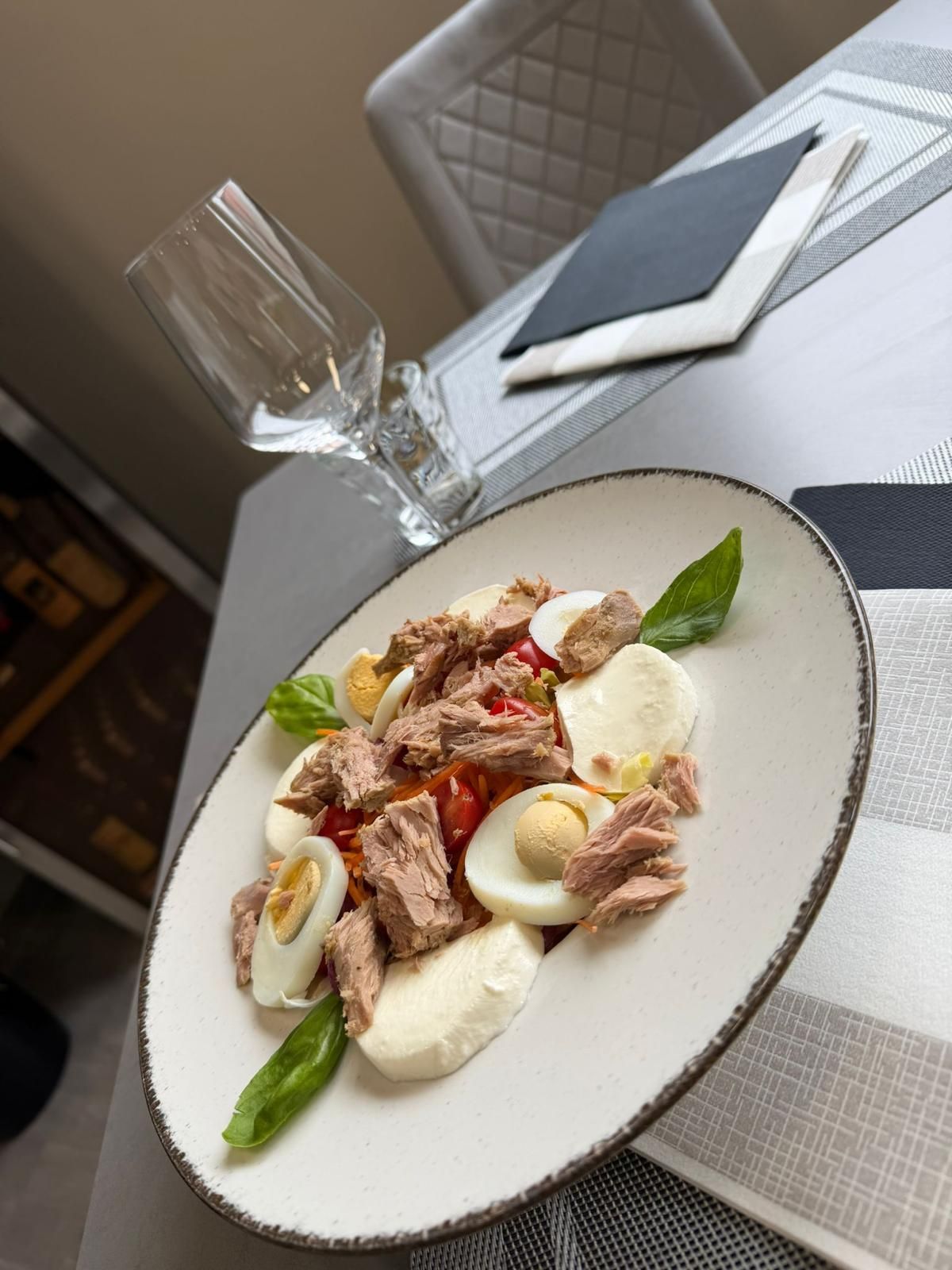 Un'insalata gourmet con uova, tonno e basilico è disposta su un tavolo accanto a un calice di vino e un tovagliolo piegato.