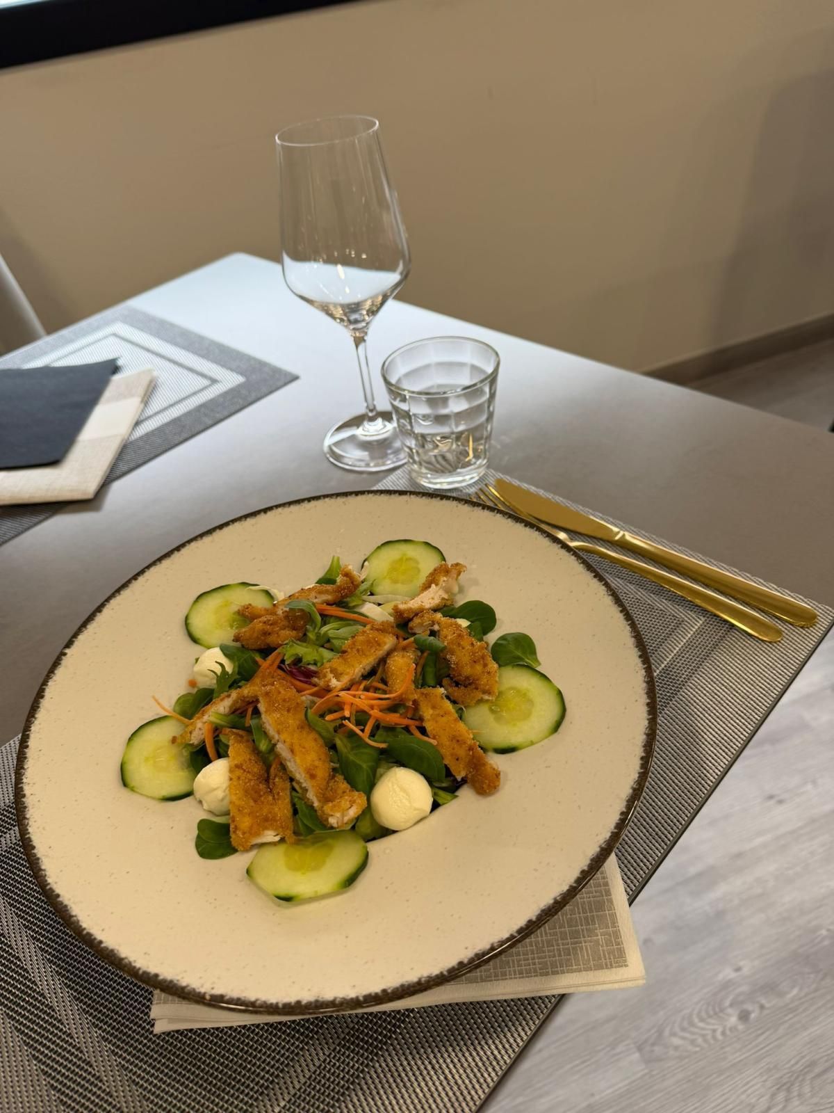 Un'insalata gourmet con cetrioli a fette e verdure a foglia verde, servita su un tavolo di ristorante accanto a bicchieri di vino e acqua.