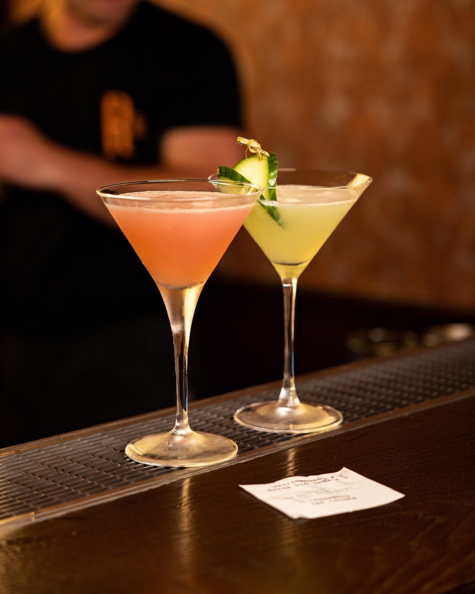 Two cocktails in martini glasses on a bar. One pink, one green, garnished with cucumber.