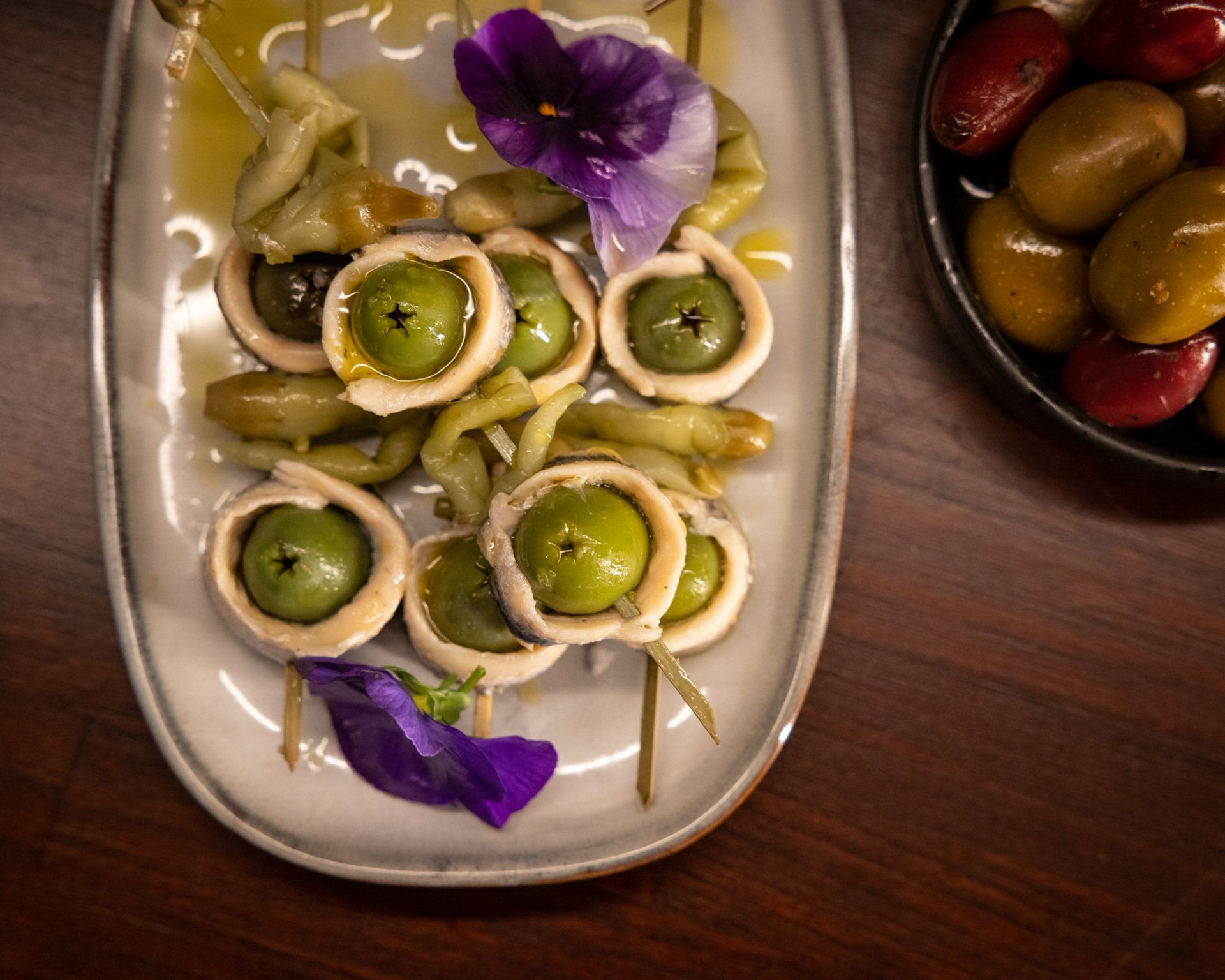 Gildas appetizer with green olives, anchovies, and peppers on a platter, next to a bowl of olives.