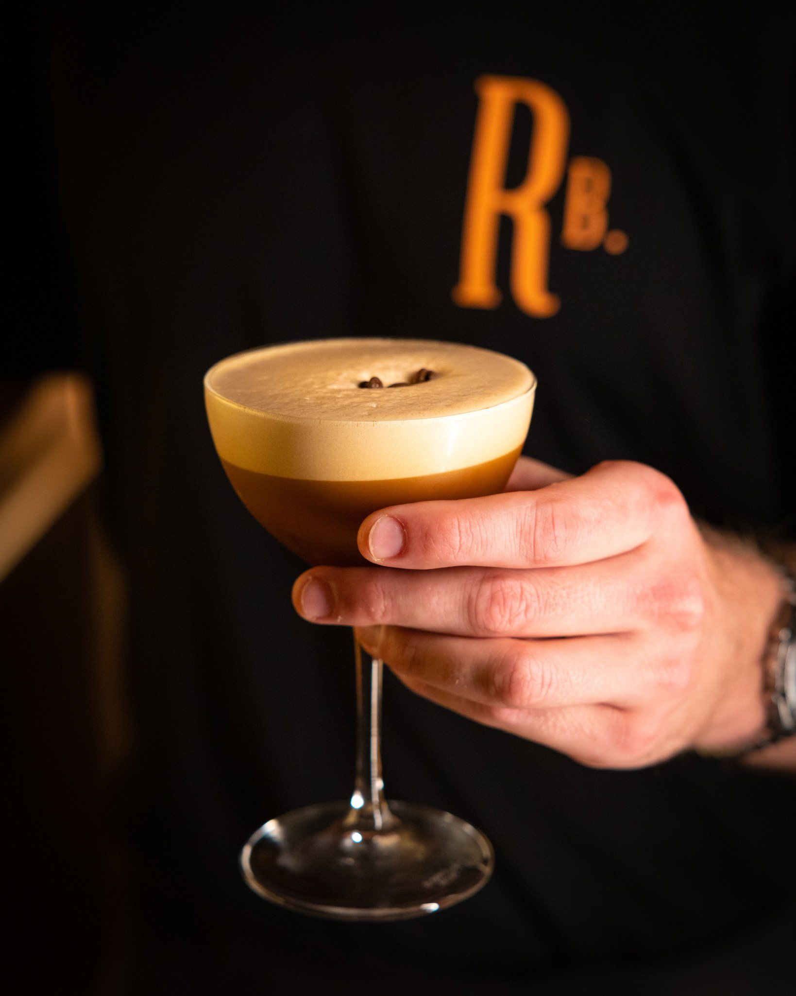 A person holding an espresso martini in a stemmed glass with a frothy top; dark background.
