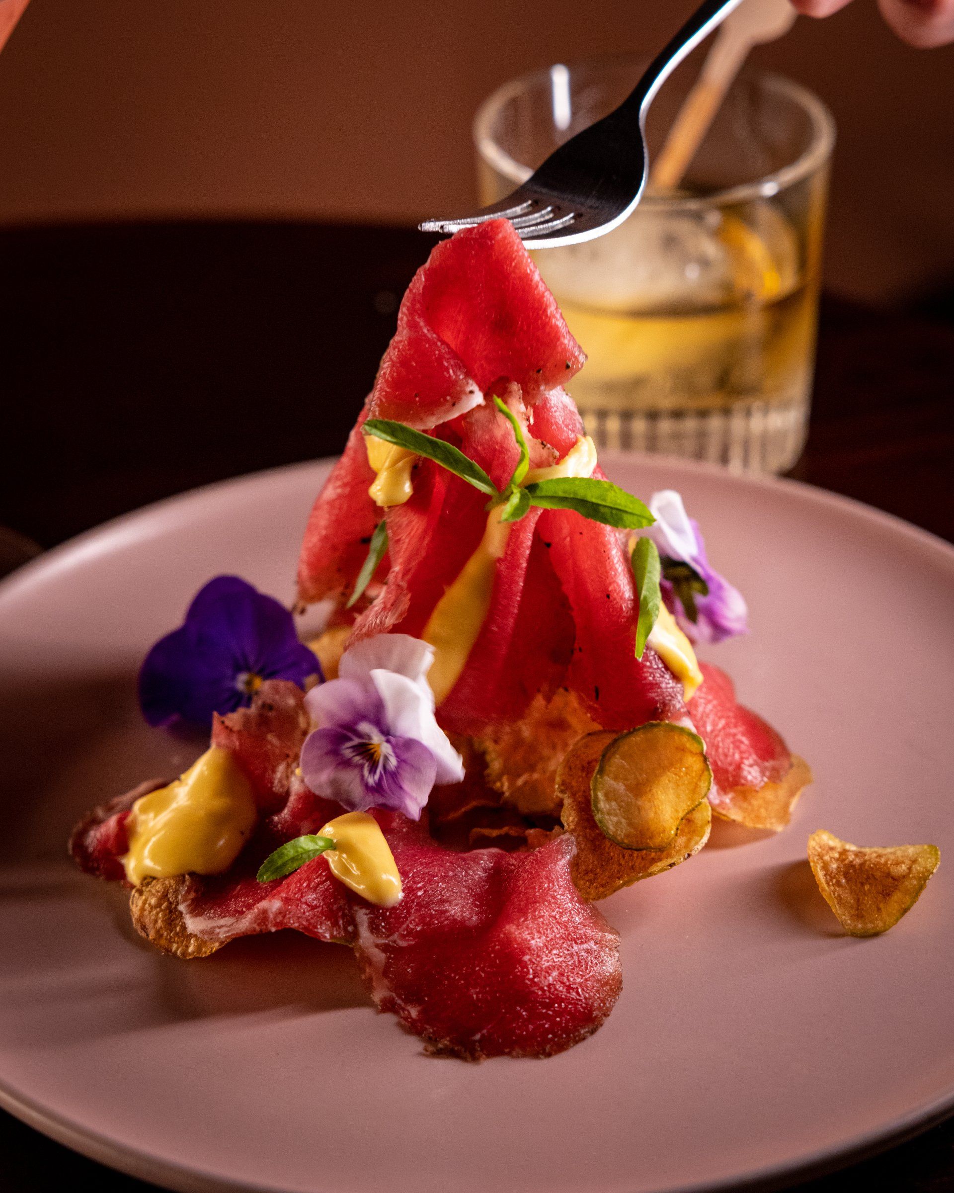 A pink plate with a gourmet dish featuring beef carpaccio, flowers, and a bearnaise sauce. A fork is poised above.