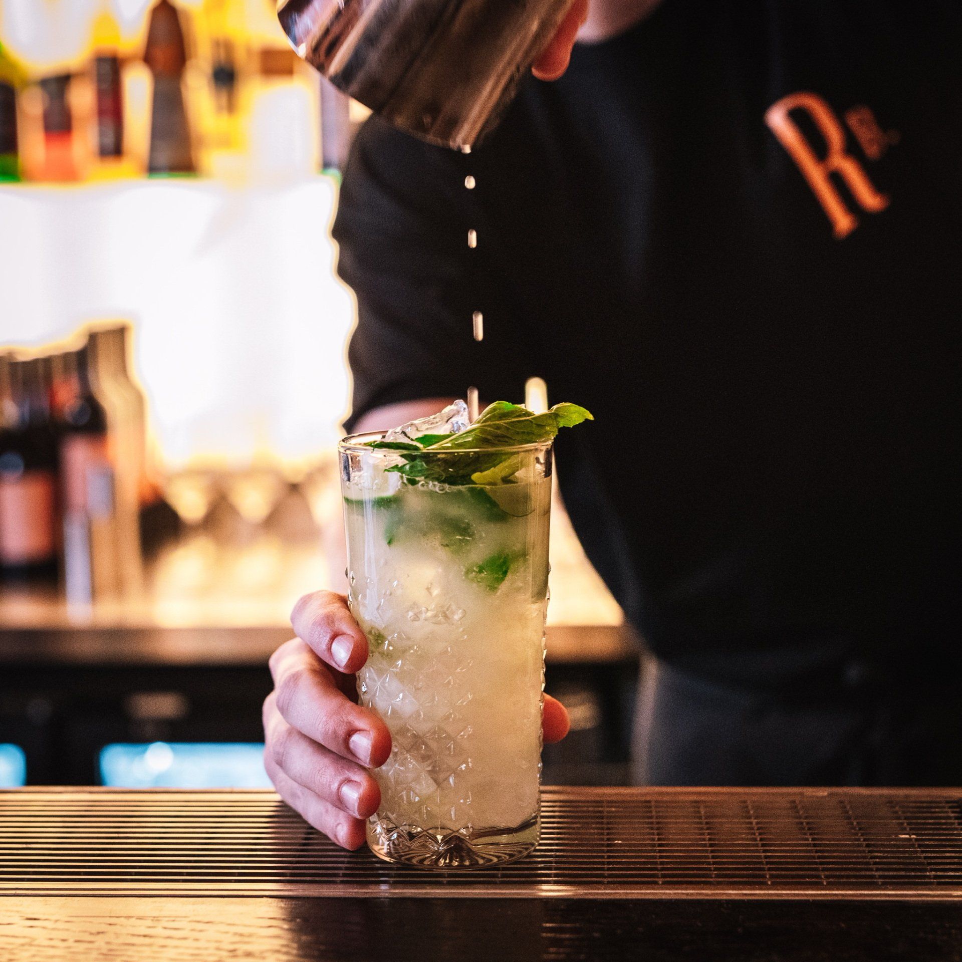 Bartender pouring a Mojito into a cocktail glass with mint, at a bar.