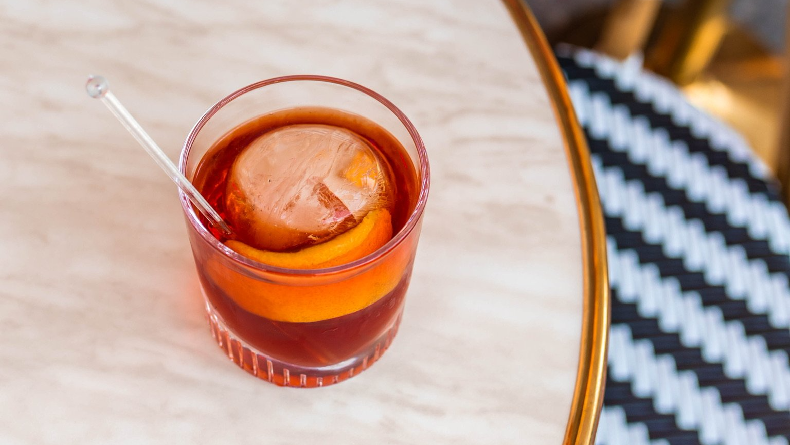 Cocktail in a glass with orange peel and a large ice sphere, on a marble table.
