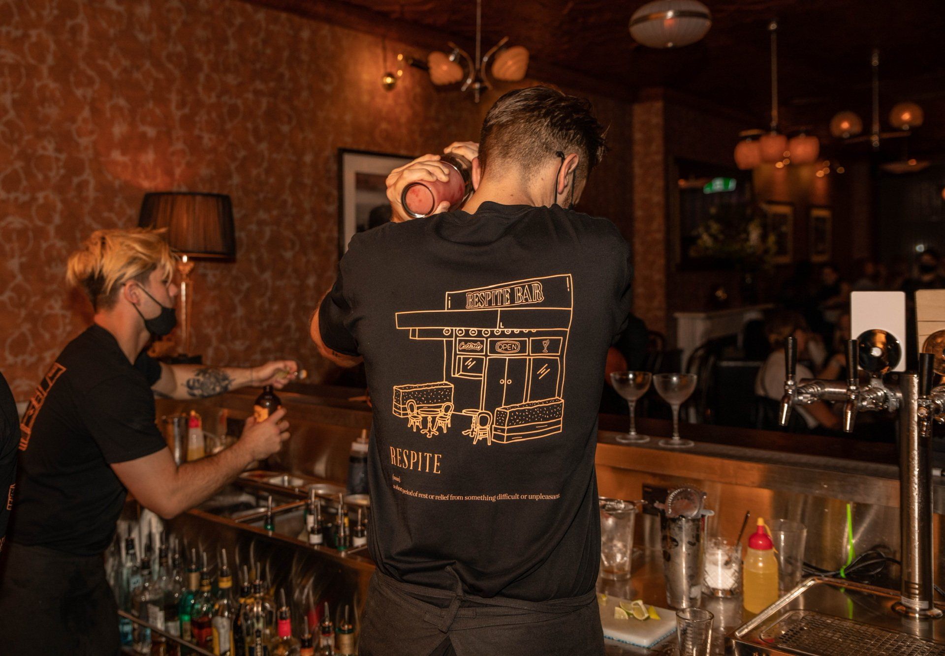 Bartenders at work in a dimly lit bar, one shaking a cocktail, the other pouring a drink.