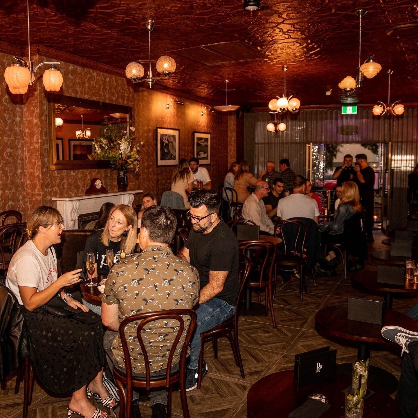 People gathered inside a moody bar, conversing at tables, under globe pendant lights.