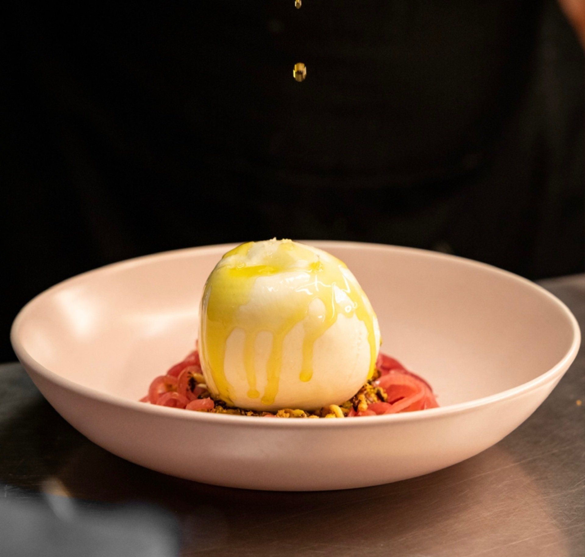 Burrata cheese topped with olive oil in a pink bowl on a metal surface.