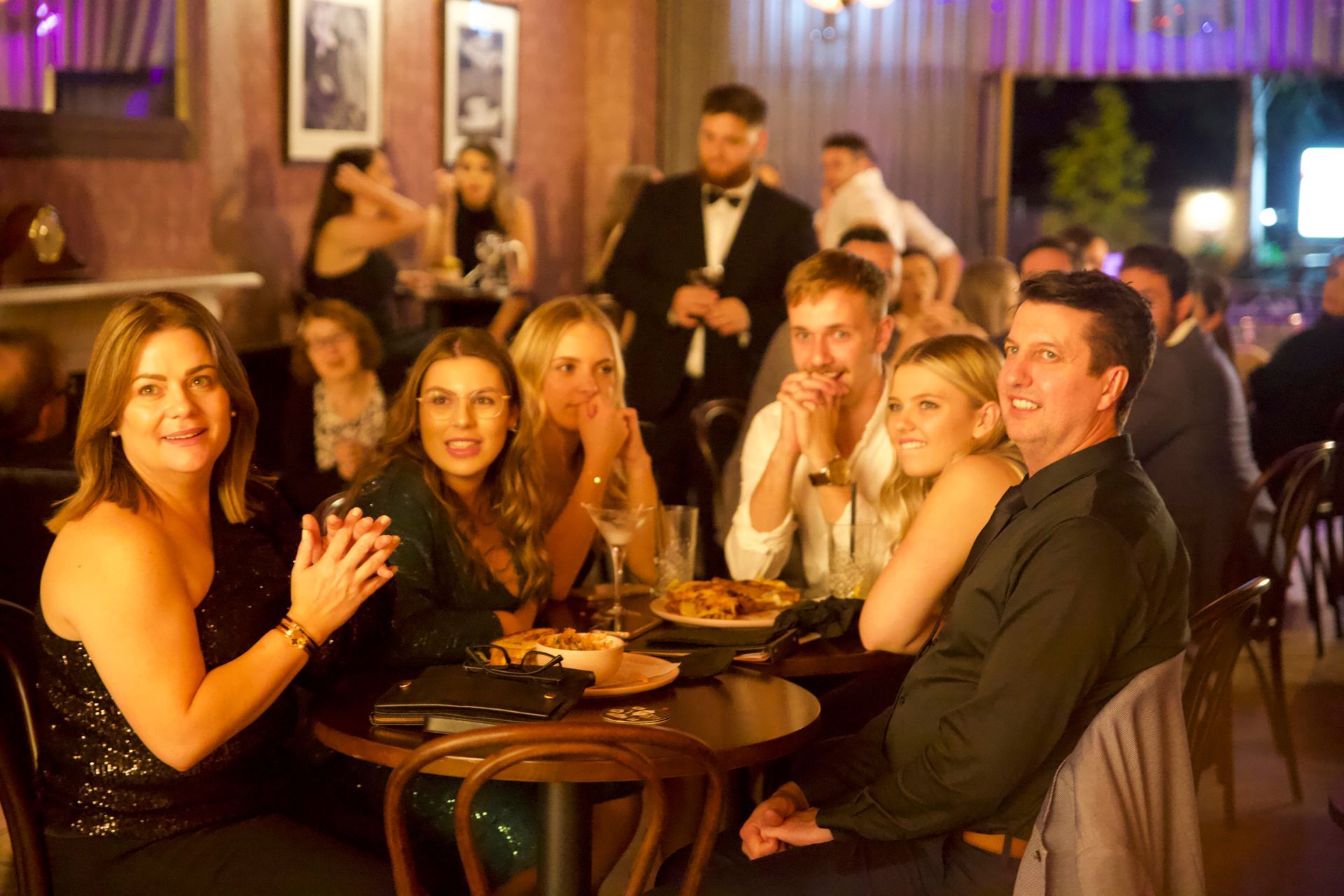 People at a dimly lit bar playing trivia; woman applauds, others watch. Wooden tables, a waiter in a suit.