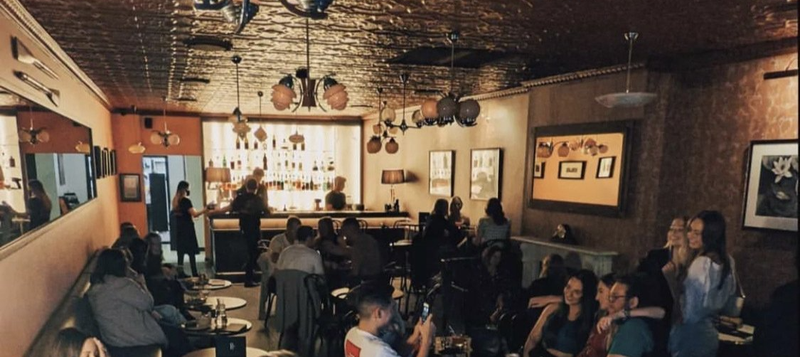 Interior of a bar, people seated at tables and the bar, dim lighting, mirrors, and artwork on walls.