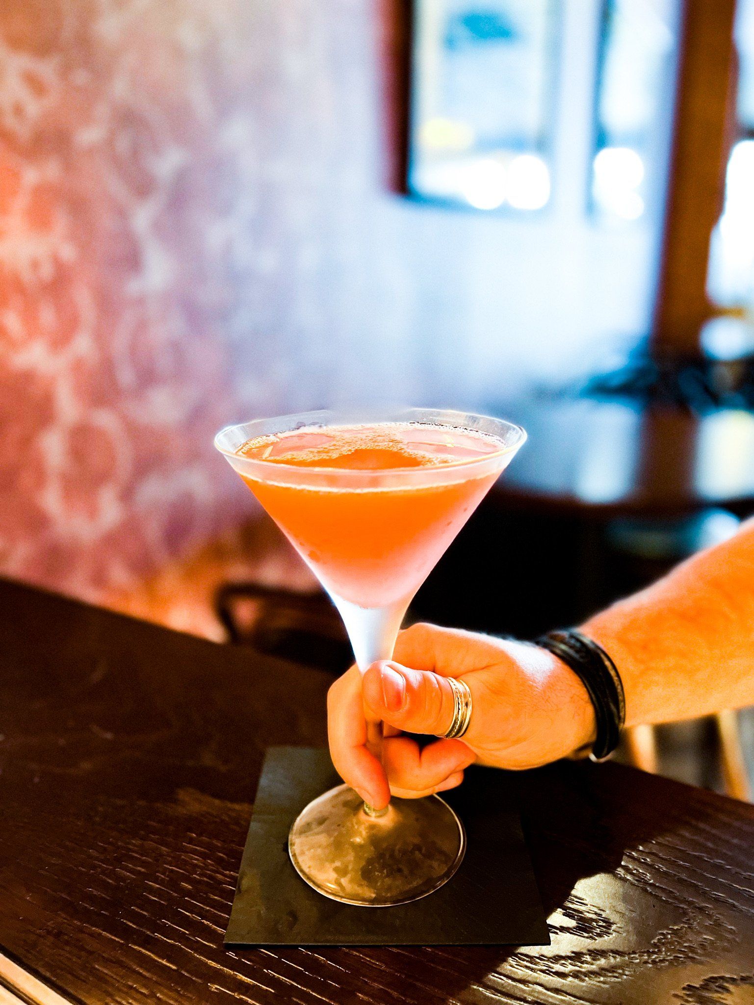 Hand holding a Cosmopolitan cocktail in a martini glass, set on a coaster on a dark bar.