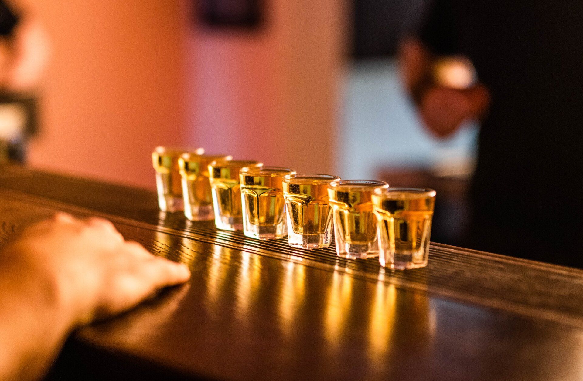 Row of seven shots on a bar, with a hand reaching towards them.