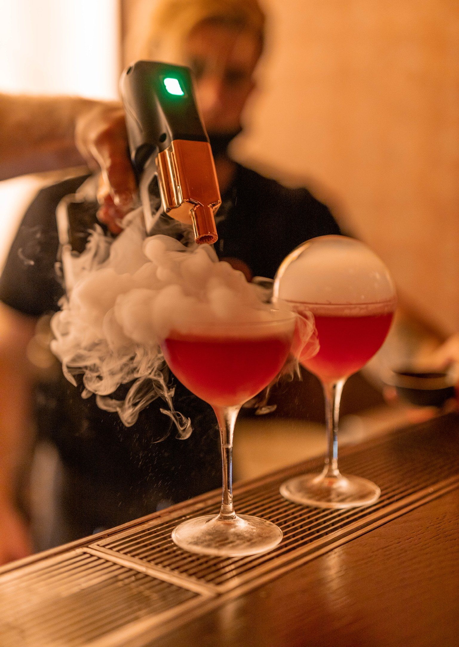 Cocktails being smoked by a bartender with a smoking gun. Smoke billows over the ruby red drinks.