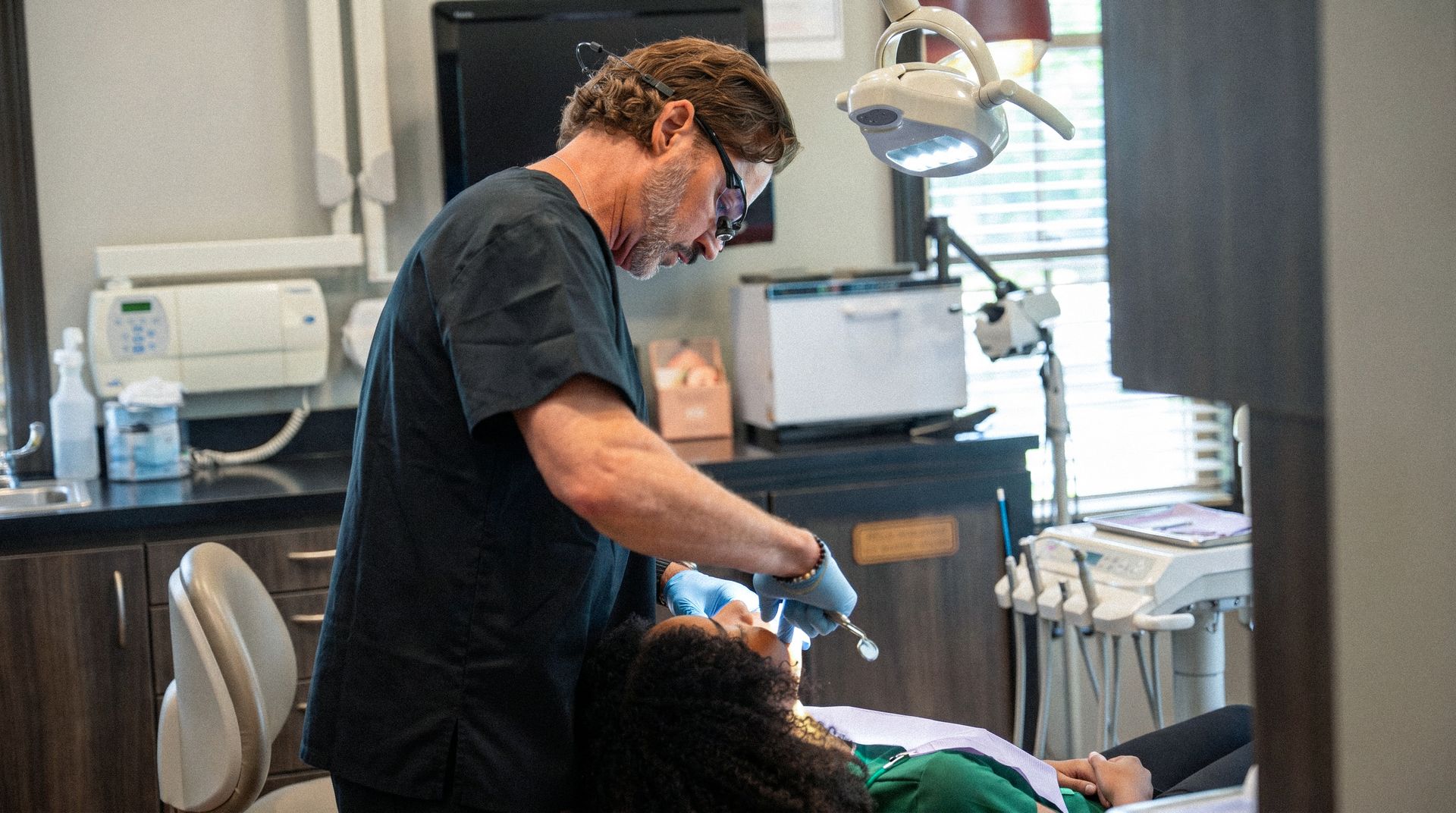 Dentist examining a patient in a clinic, wearing gloves and a face shield, with dental equipment nearby