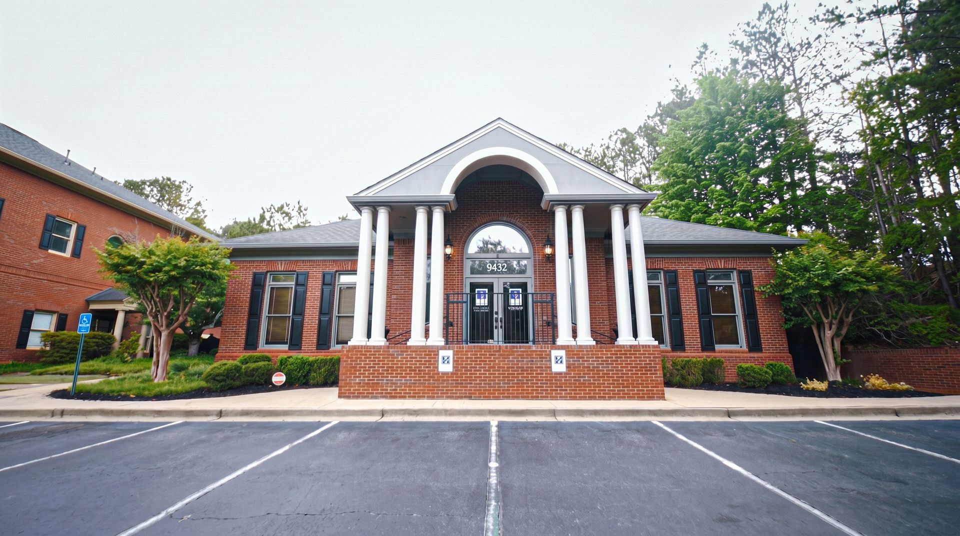 Brick building with white columns and red doors, centered in an empty parking lot under cloudy skies