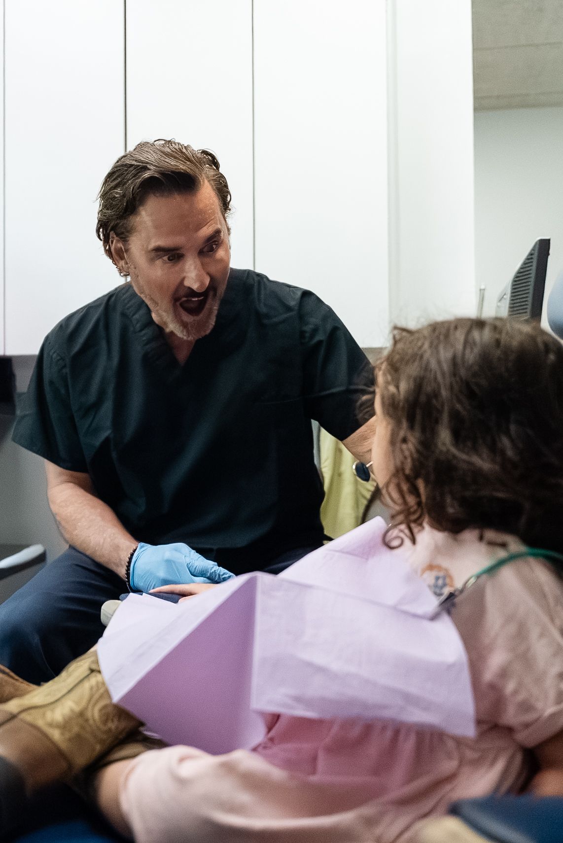 Dentist speaking with patient in exam chair, wearing blue gloves and pink bib in a clinic room