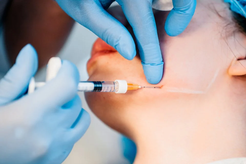 Gloved hands give an injection in a person’s neck or jaw area with a syringe.