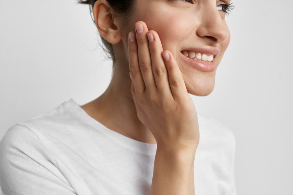 Smiling woman in a white shirt touching her cheek against a light gray background