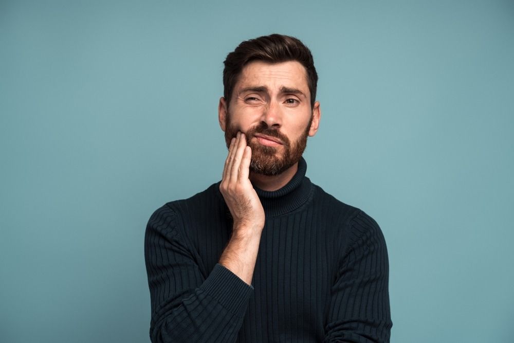 Man in a black sweater touching his cheek, looking concerned against a blue background