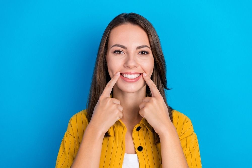 Smiling woman pointing to her teeth against a blue background, wearing a yellow shirt
