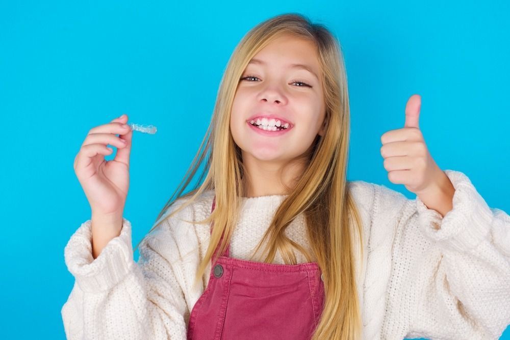 Smiling child in a white sweater and pink overalls giving a thumbs-up against a blue background