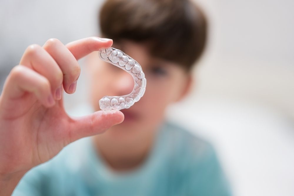 Hand holding a clear dental aligner in front of a blurred child in a blue shirt