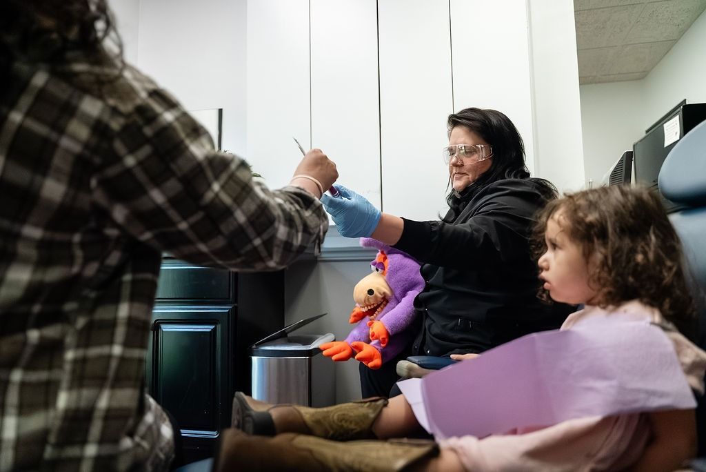Adult speaking to child in an office, with a seated child holding a purple paper and a doll nearby.