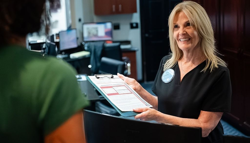Woman at an office desk smiling and holding a clipboard in a reception area