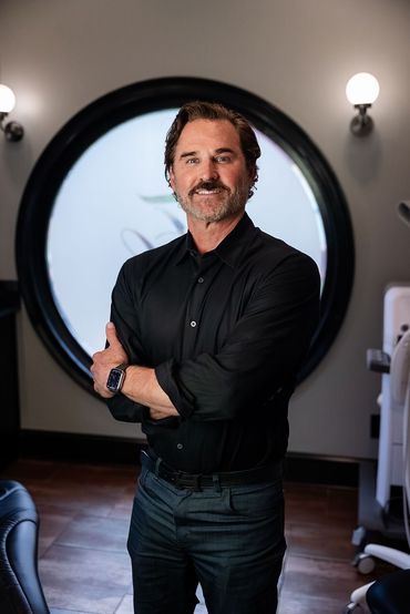 Man standing with arms crossed in a black shirt, in front of a round window and wall lights.