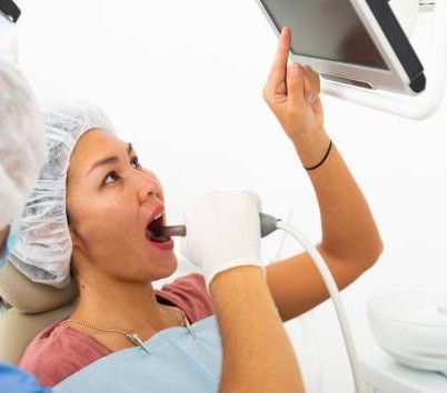 Woman in a dental chair holding a tablet while a dentist examines her mouth with a lighted tool.