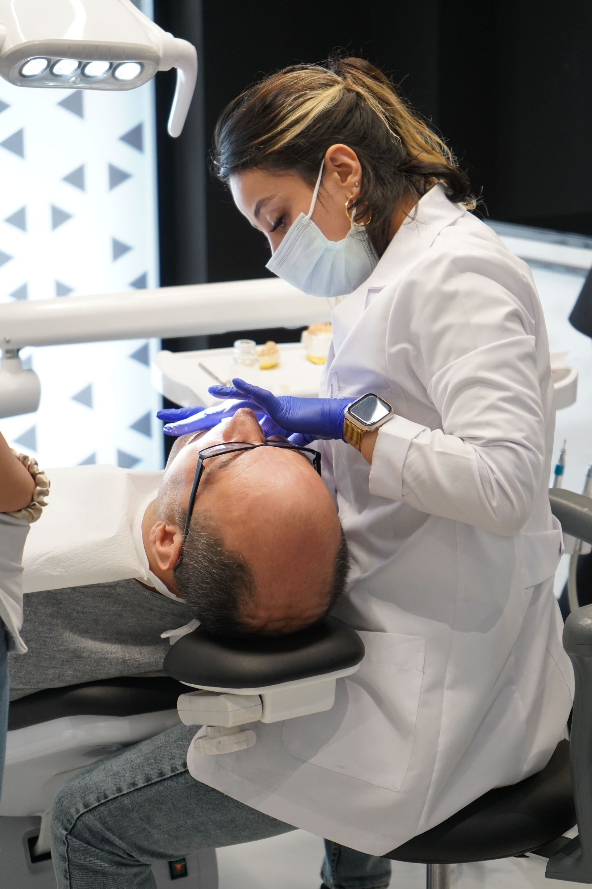 Dentist in white coat and mask examining a patient reclined in a dental chair under bright lights.
