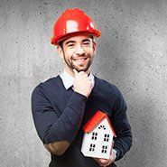 Man in hard hat holding a house model, smiling, posing in front of a gray wall.
