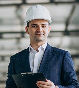 Man in a hard hat and suit holding a clipboard, indoors.