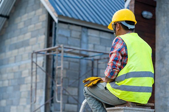 Construction worker wearing yellow hard hat and vest sits, looking at building under construction.