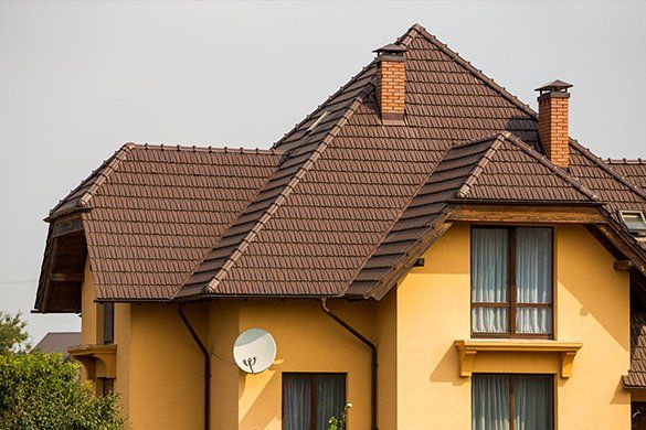Yellow house with brown tile roof and two chimneys, against a bright sky.