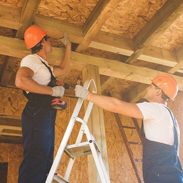 Two construction workers in orange helmets install wooden beams.